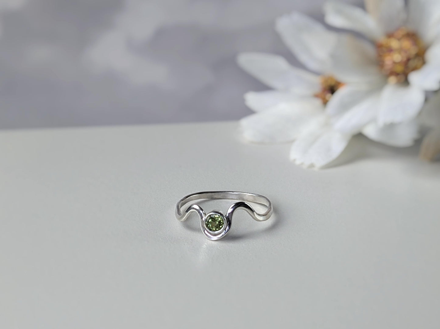 Slightly angled view of a ring. The ring is made from sterling silver. It has a wave design, with a peridot set in a bezel in the middle of the waves.