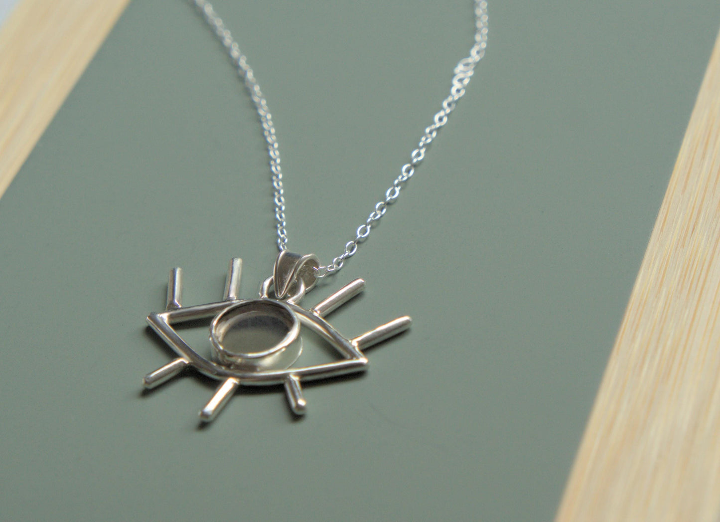 An angled view of a pendant. It is an eye-shaped pendant sitting on a simple chain. The pendant is mader from sterling silver and has eyelashes made of silver wire and an iris made of smokey quartz.