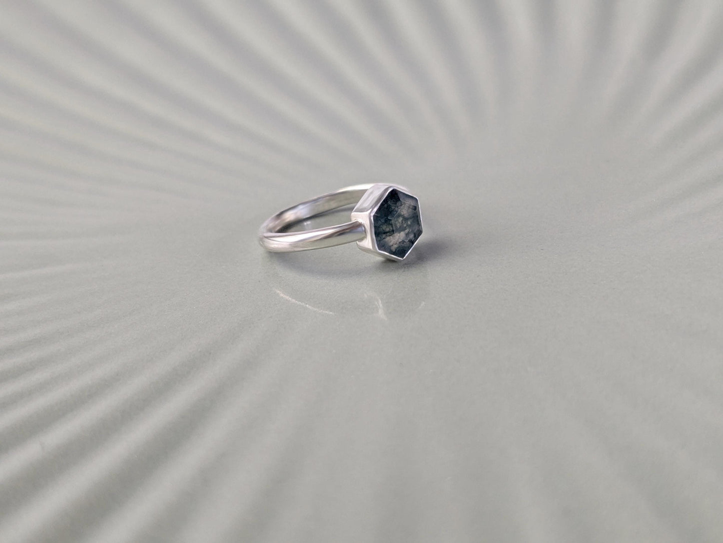 An angled view of a ring. The ring is made from sterling silver. The ring has a plain band and a hexagon-shaped bezel. The bezel holds a faceted moss agate.