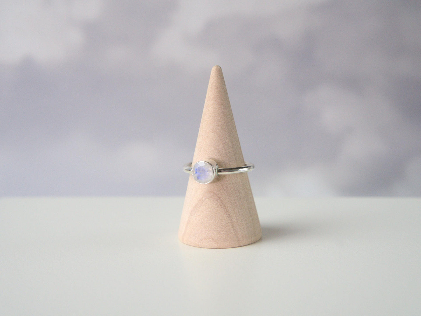 A side view of a ring. The ring has a plain sterling silver band with a round bezel. The bezel holds faceted moonstone.
