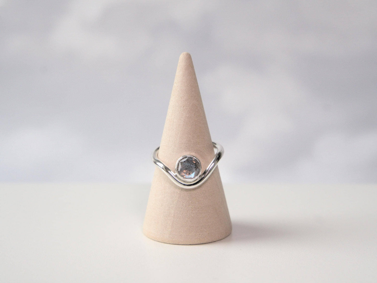 A front-on view of a ring sitting on a ring cone. The ring is made from sterling silver and has a band that curves downwards in the front. In the trough of the curve sits a round bezel with a faceted labradorite gemstone.