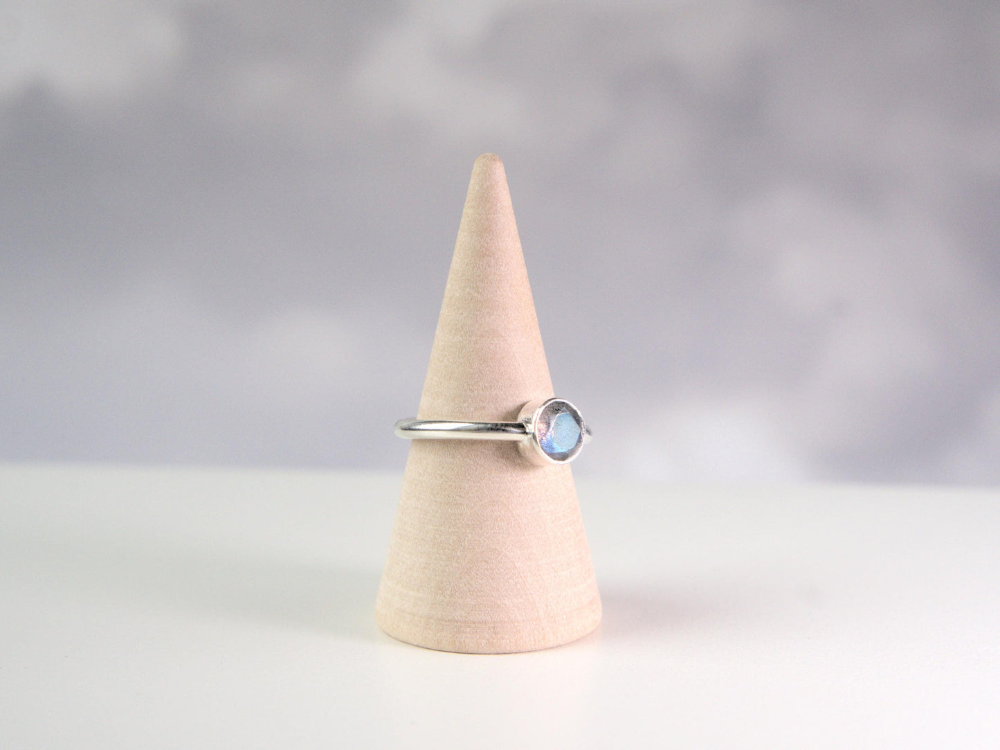 A side view of a ring. It is a simple ring displayed on a wooden ring cone. The ring has a plain sterling silver band with a round bezel, which holds a faceted labradorite gemstone.