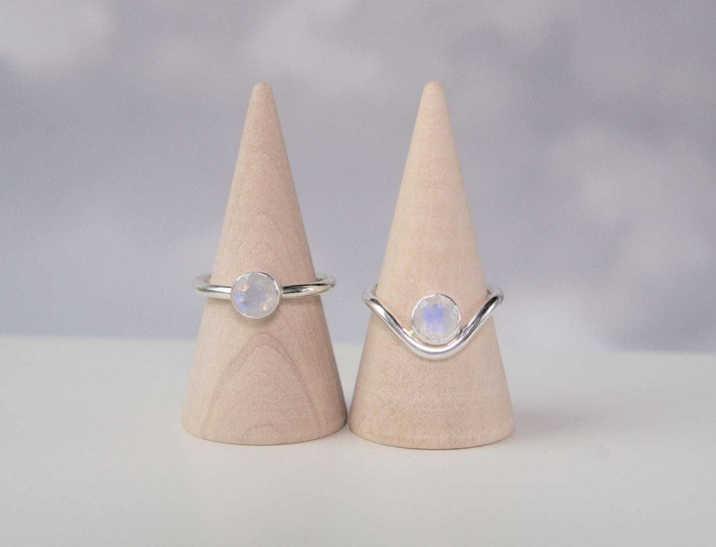 A comparison of two rings. Both are made from sterling silver and have a round bezel that holds a faceted moonstone. The ring on the left has a plain band. The ring on the right has a band which curves downwards in the front. The ring with the curved band has the gemstone nestled within the trough of the curve.