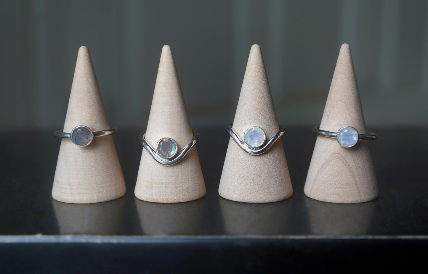 A comparison of different rings, displayed on ring cones. The two rings on the left have labradorite gemstones. The two rings on the right have moonstones. The rings show the plain and curved variants of band with each gemstone.