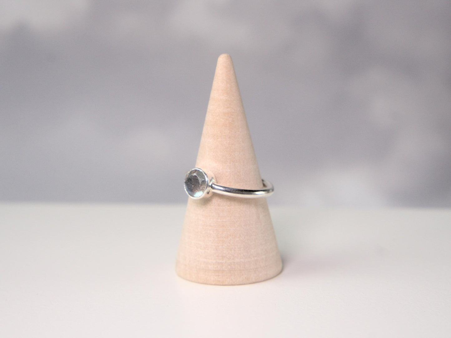 A side view of a ring. It is a simple ring displayed on a wooden ring cone. The ring has a plain sterling silver band with a round bezel, which holds a faceted labradorite gemstone.