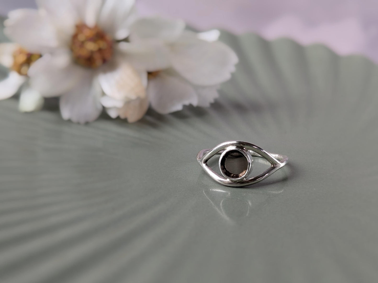 A simple sterling silver ring in the shape of an eye. The iris is made of a transparent disc of smokey quartz. There is an out-of-focus bunch of flowers in the background.