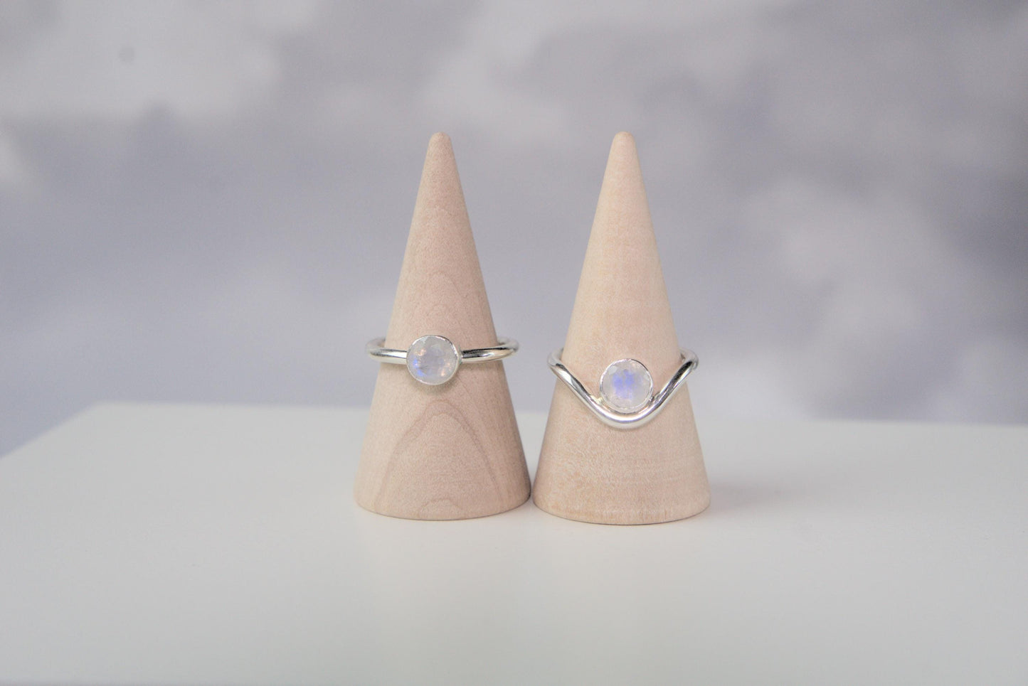 A comparison of two rings. Both are made from sterling silver and have a round bezel that holds a faceted moonstone. The ring on the left has a plain band. The ring on the right has a band which curves downwards in the front. The ring with the curved band has the gemstone nestled within the trough of the curve.
