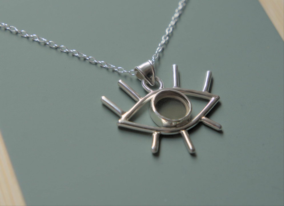 An angled view of a pendant. It is an eye shaped pendant sitting on a simple chain. The pendant is mader from sterling silver and has eyelashes made of silver wire and an iris made of smokey quartz.