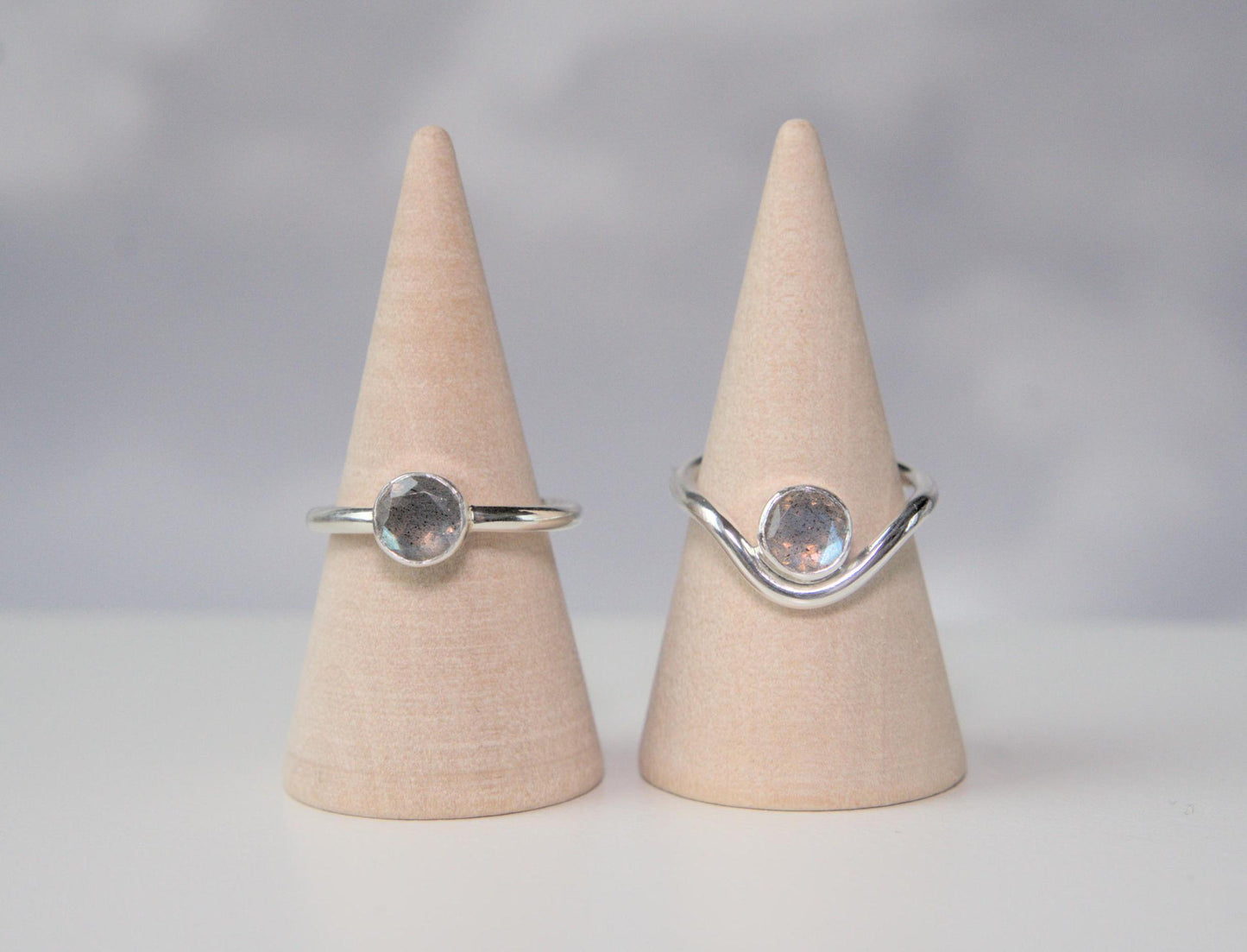 A comparison of two rings. Both are made from sterling silver and have a round bezel that holds a faceted labradorite. The ring on the left has a plain band. The ring on the right has a band which curves downwards in the front. The ring with the curved band has the gemstone nestled within the trough of the curve.