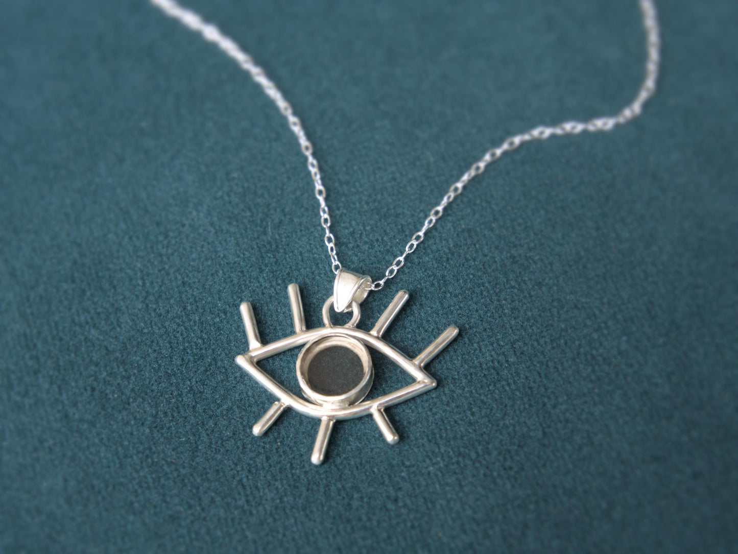 An angled view of a pendant. It is an eye shaped pendant sitting on a simple chain. The pendant is mader from sterling silver and has eyelashes made of silver wire and an iris made of smokey quartz.