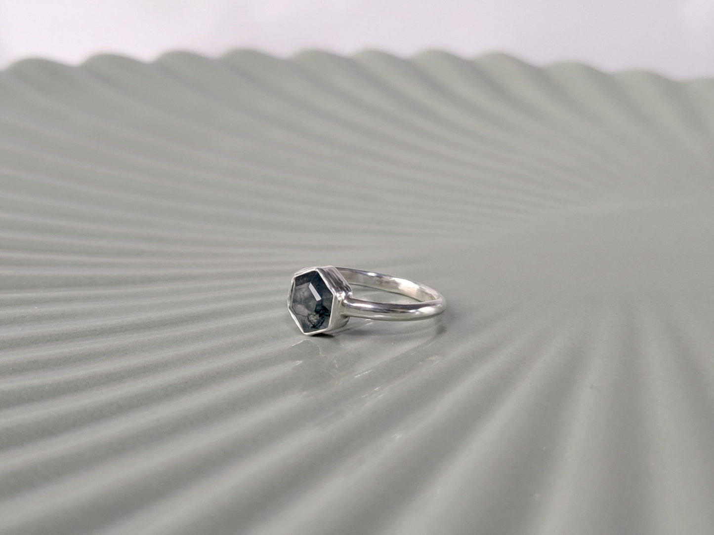 An angled view of a ring. The ring is made from sterling silver. The ring has a plain band and a hexagon-shaped bezel. The bezel holds a faceted moss agate.