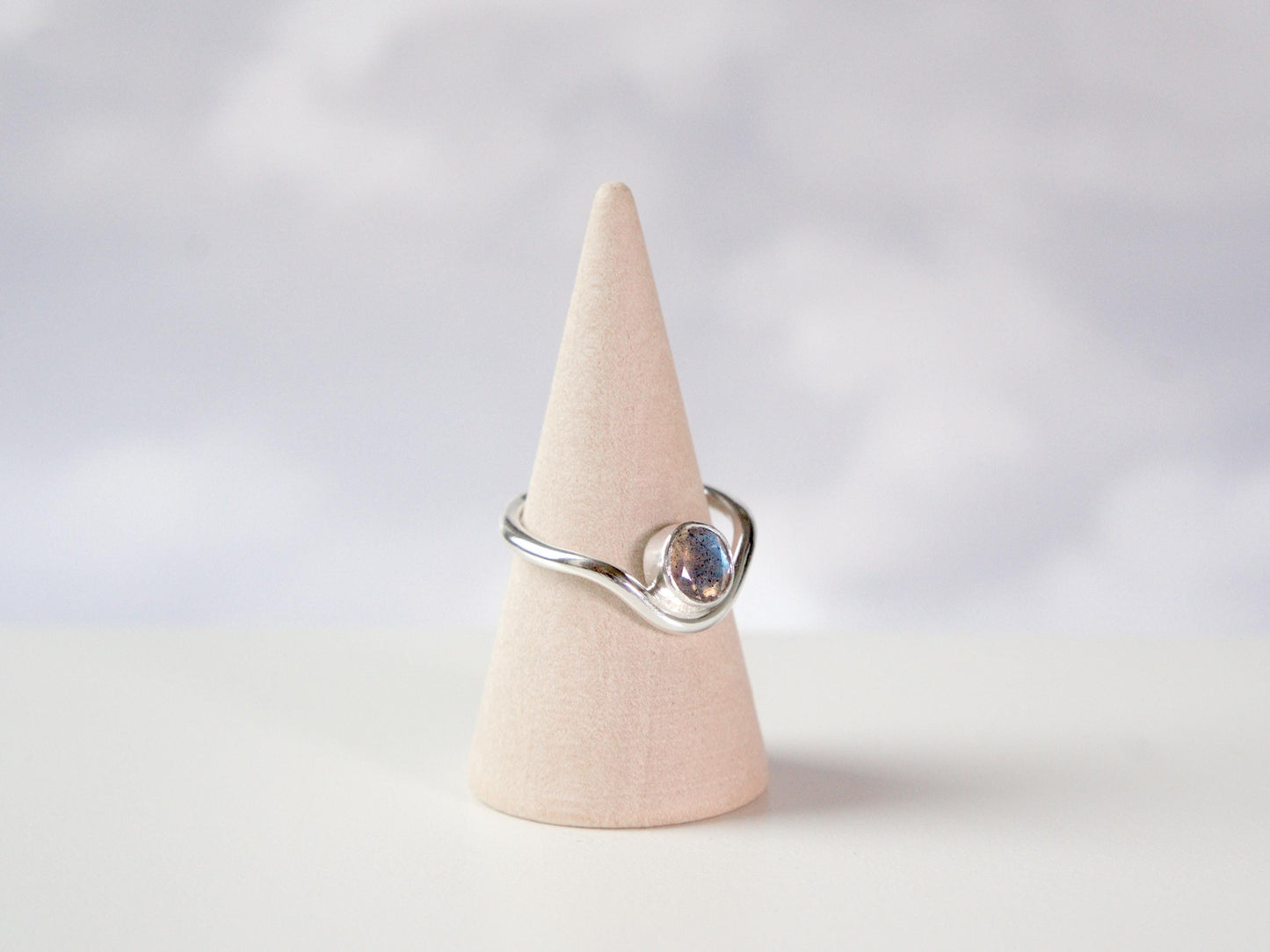 A side view of a ring sitting on a ring cone. The ring is made from sterling silver and has a band that curves downwards in the front. In the trough of the curve sits a round bezel with a faceted labradorite gemstone.