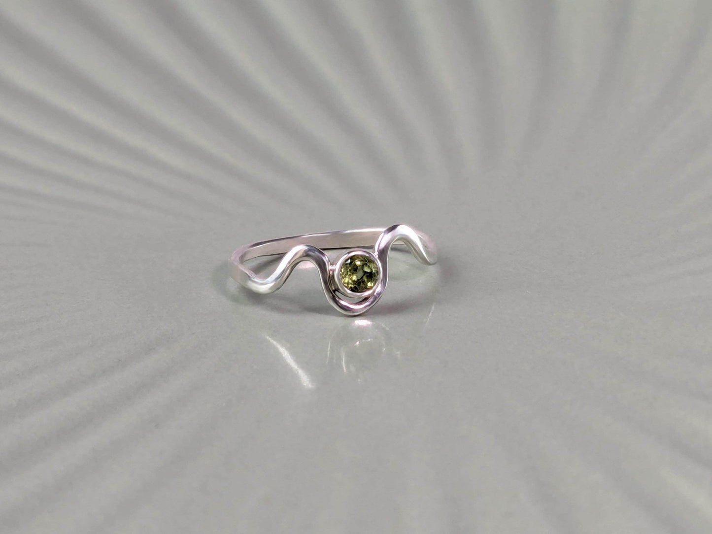 Frontal view of a ring. The ring is made from sterling silver. It has a wave design, with a peridot set in a bezel in the middle of the waves.