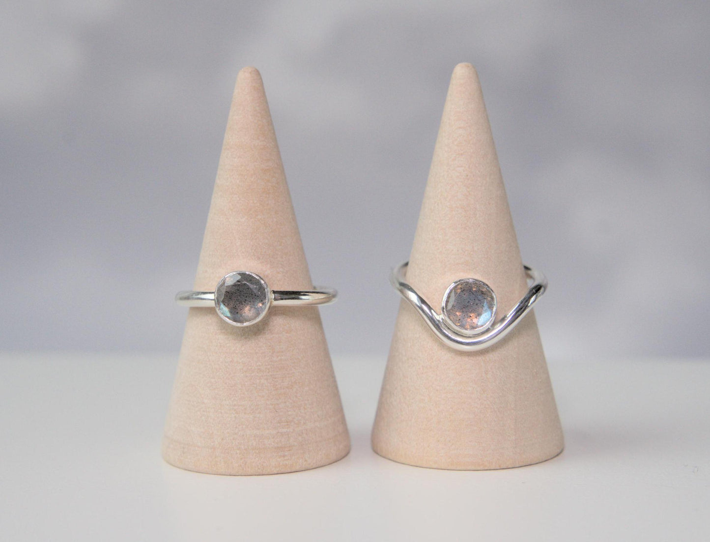 A comparison of two rings. Both are made from sterling silver and have a round bezel that holds a faceted labradorite. The ring on the left has a plain band. The ring on the right has a band which curves downwards in the front. The ring with the curved band has the gemstone nestled within the trough of the curve.