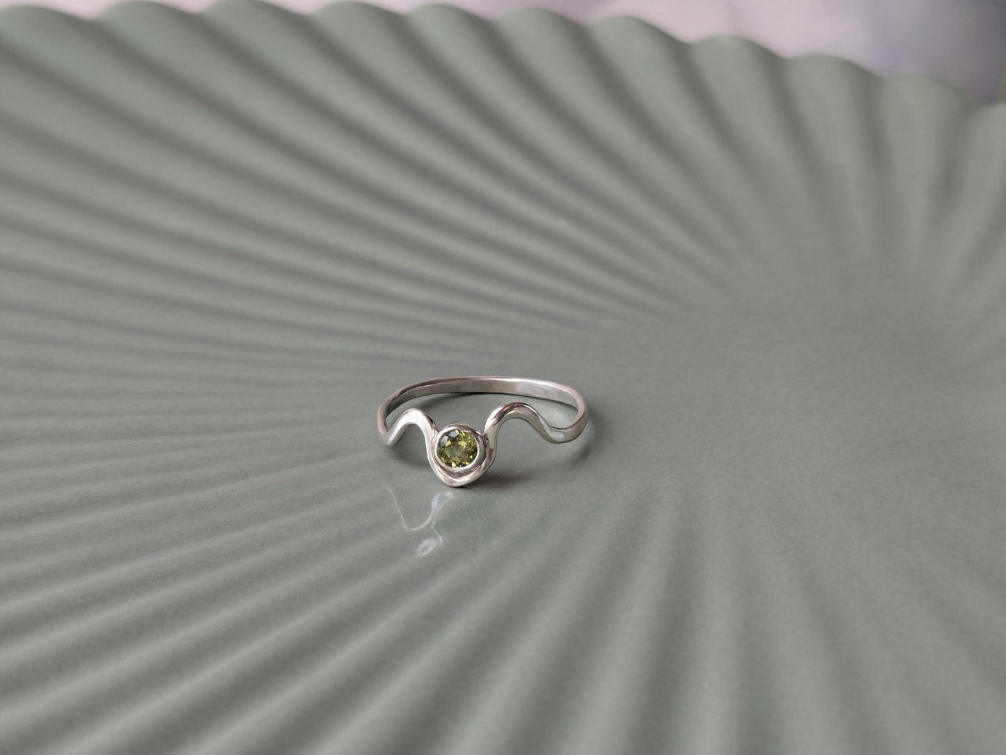 Angled view of a ring. The ring is made from sterling silver. It has a wave design, with a peridot set in a bezel in the middle of the waves. 