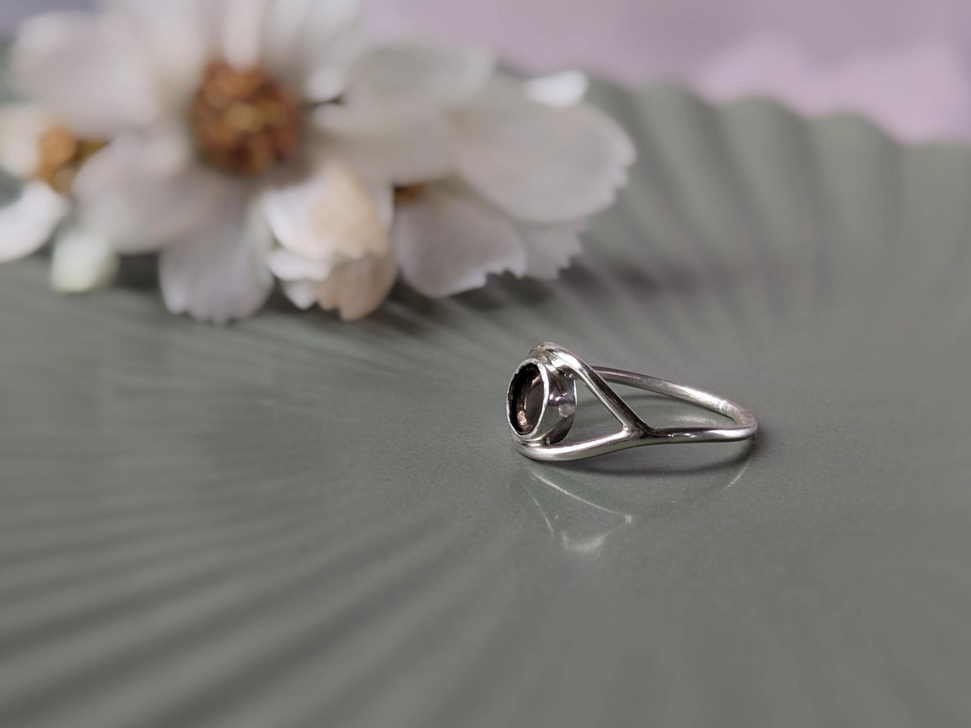 A side view of a ring. The ring is made from sterling silver ring and forms the shape of an eye. The iris is made of a transparent disc of smokey quartz. There is an out-of-focus bunch of flowers in the background.