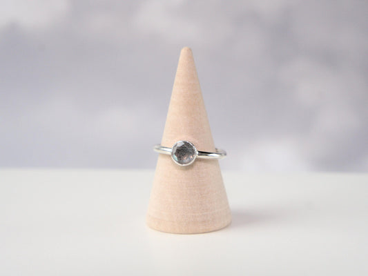 A frontal view of a ring. It is a simple ring displayed on a wooden ring cone. The ring has a plain sterling silver band with a round bezel, which holds a faceted labradorite gemstone. 