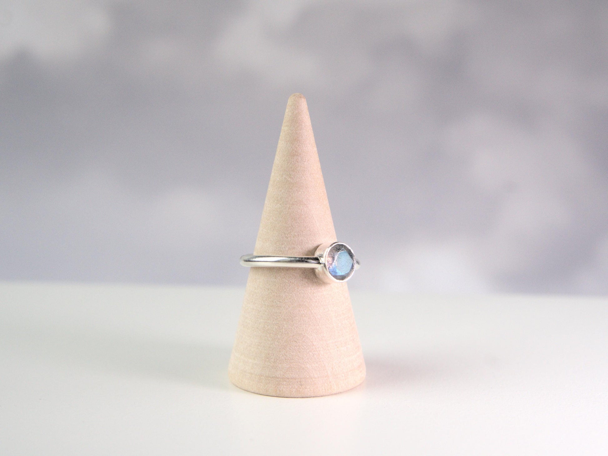 A side view of a ring. It is a simple ring displayed on a wooden ring cone. The ring has a plain sterling silver band with a round bezel, which holds a faceted labradorite gemstone. 
