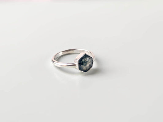 An angled view of a ring with a plain background. The ring is made from sterling silver. The ring has a plain band and a hexagon-shaped bezel. The bezel holds a faceted moss agate.
