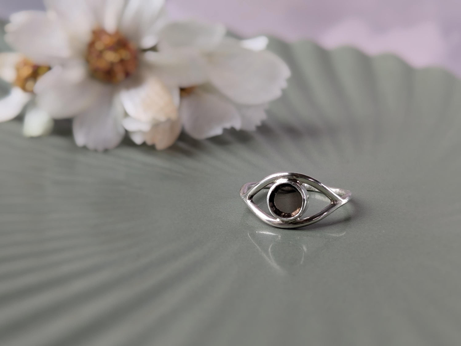 A simple sterling silver ring in the shape of an eye. The iris is made of a transparent disc of smokey quartz. There is an out-of-focus bunch of flowers in the background.