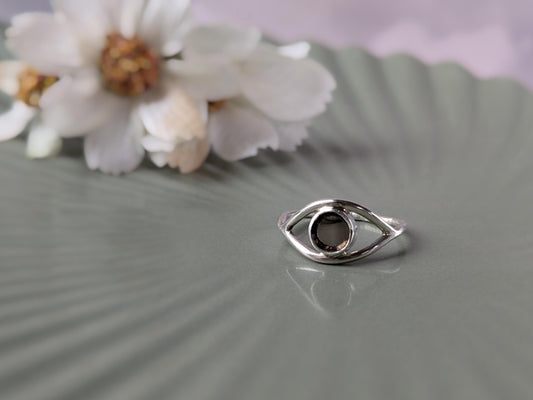 A simple sterling silver ring in the shape of an eye. The iris is made of a transparent disc of smokey quartz. There is an out-of-focus bunch of flowers in the background.