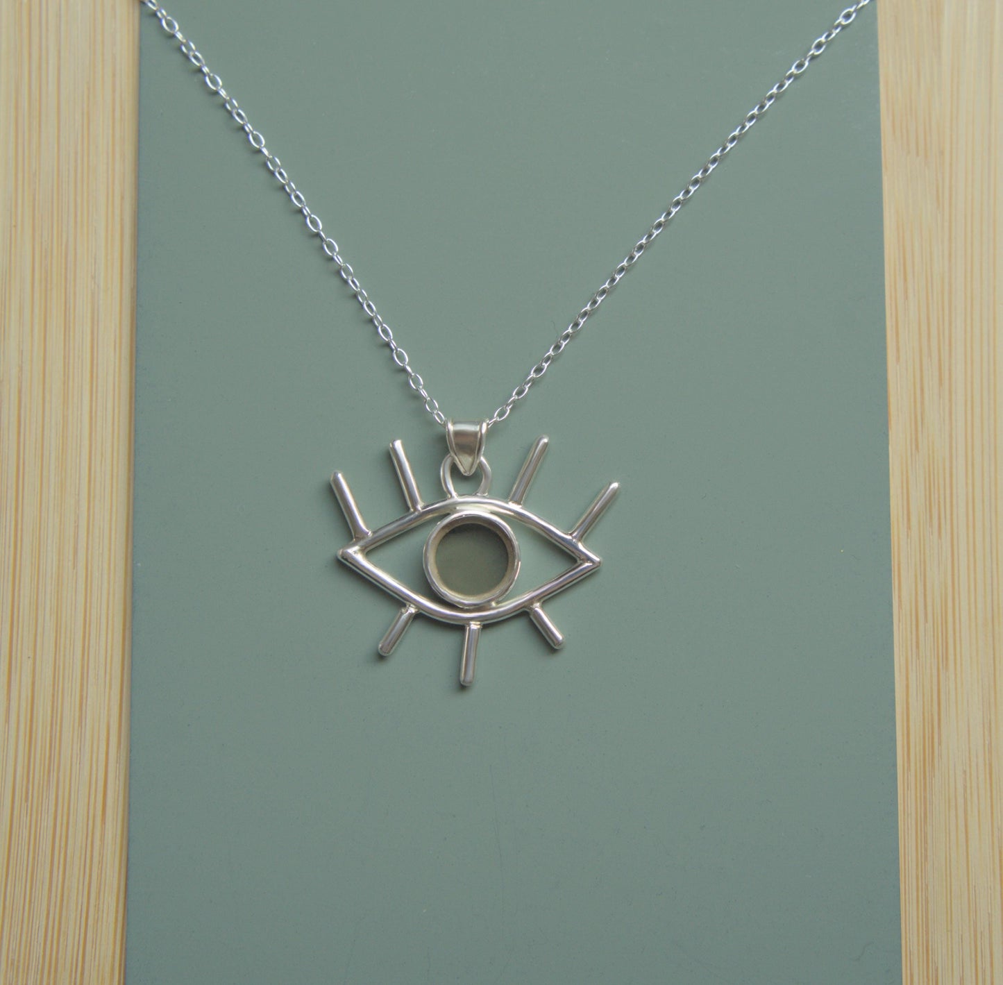 An eye-shaped pendant sitting on a simple chain. The pendant is mader from sterling silver and has eyelashes made of silver wire and an iris made of smokey quartz. 