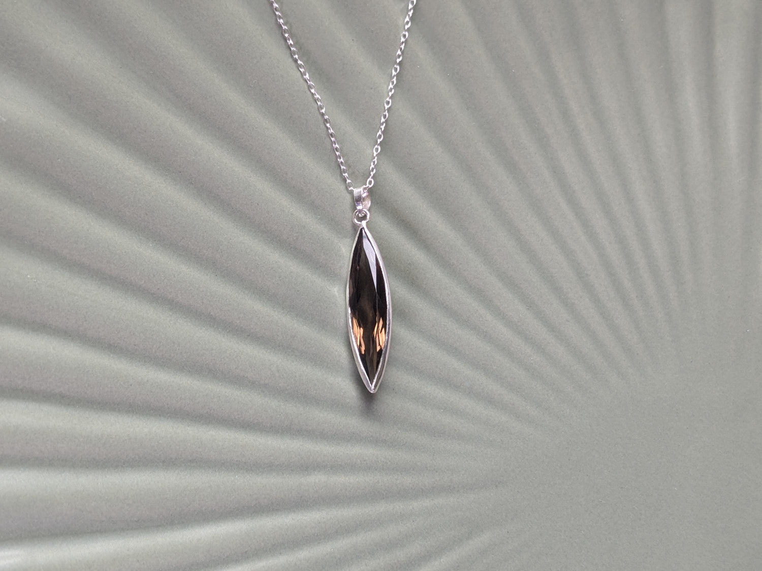 An elegant sterling silver pendant on a simple chain. The pendant is made of an elongated and slender marquise-shaped smoky quartz in a delicate bezel. 