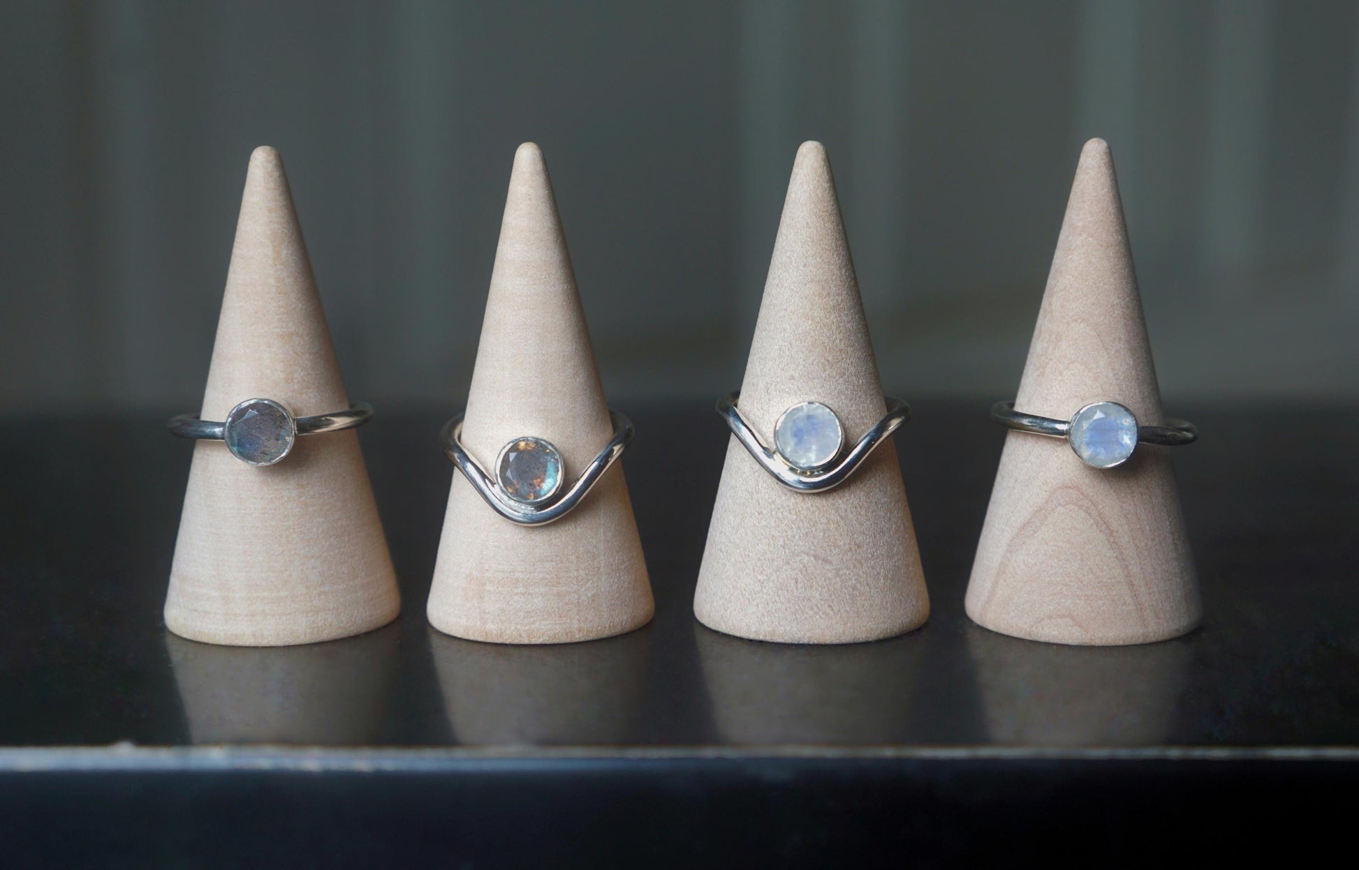 A comparison of four different rings, displayed on ring cones. The two rings on the left have labradorite gemstones. The two rings on the right have moonstones. The rings show the plain and curved variants of band with each gemstone. 