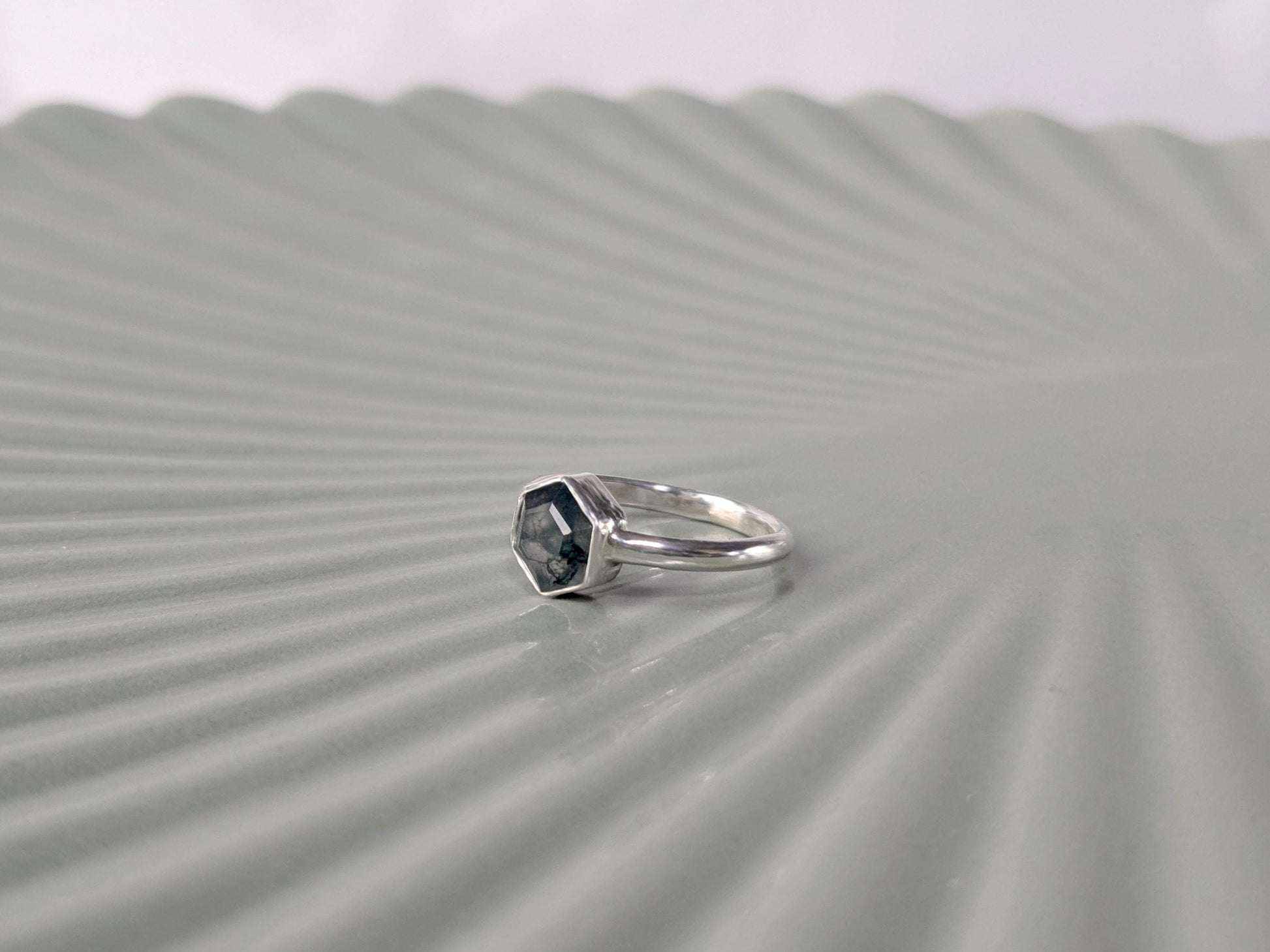 An angled view of a ring. The ring is made from sterling silver. The ring has a plain band and a hexagon-shaped bezel. The bezel holds a faceted moss agate.