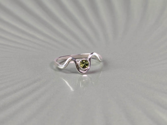 Frontal view of a ring. The ring is made from sterling silver. It has a wave design, with a peridot set in a bezel in the middle of the waves. 