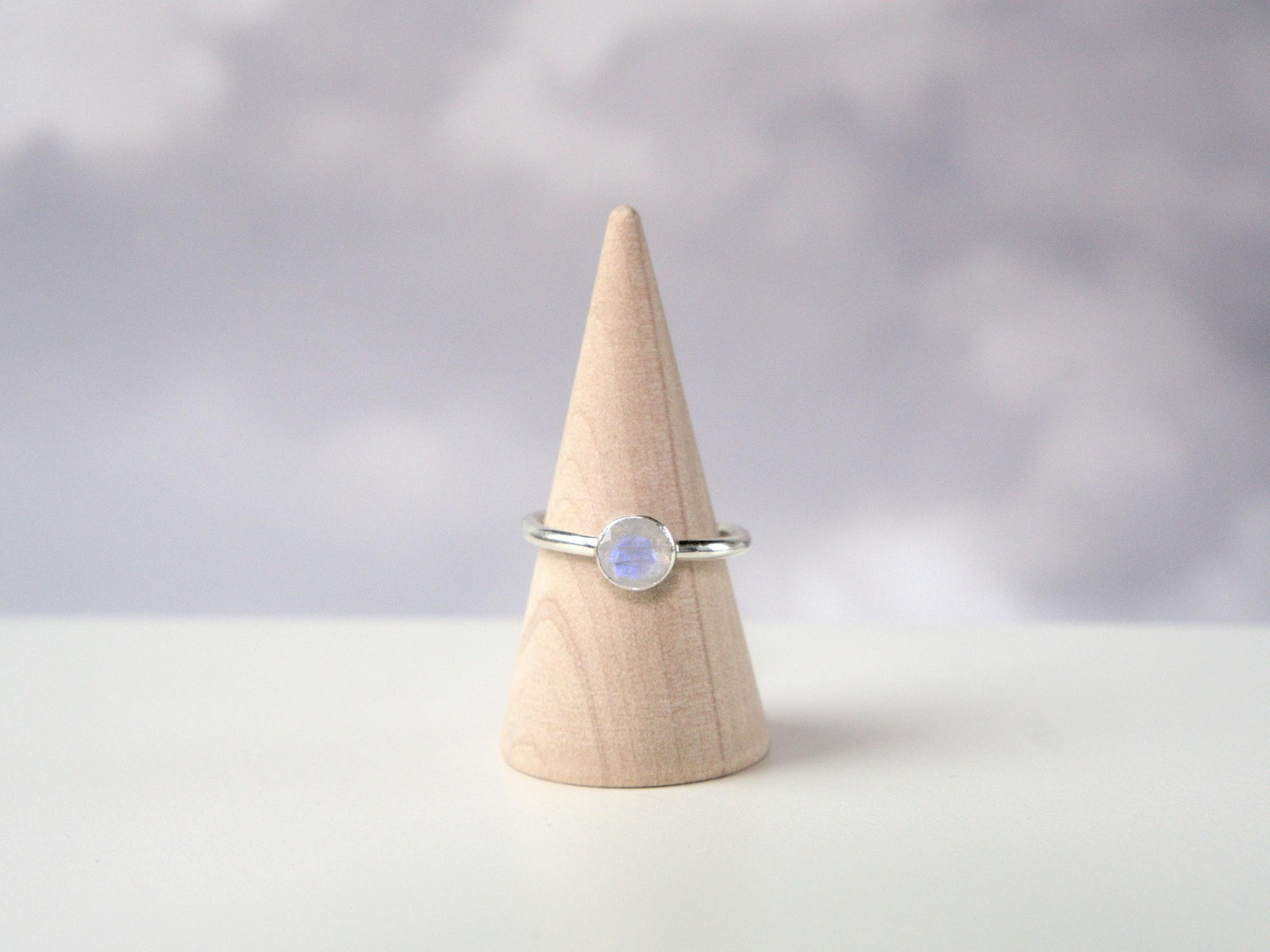 A frontal view of a ring. The ring has a plain sterling silver band with a round bezel. The bezel holds a faceted moonstone. 
